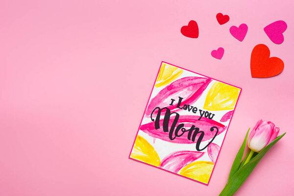 Вид сверху поздравительной открытки "Я люблю тебя, мама", лежащей рядом с тюльпанами и бумажными сердечками на розовом фоне
