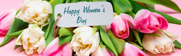 Panoramic shot of happy womens day lettering on paper label on bouquet of tulips on pink background