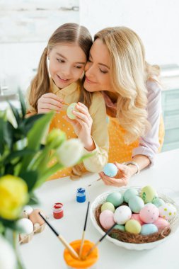 selective focus of happy mother near cute daughter painting easter egg 