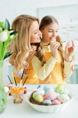 selective focus of cheerful kid painting easter egg near mother