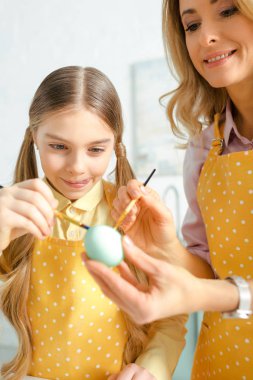 selective focus of cheerful kid sticking out tongue and painting easter egg with mother