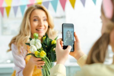 selective focus of kid taking photo of happy mother with tulips 