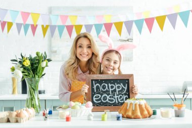 cheerful mother near tulips and daughter with bunny ears holding chalkboard with happy easter lettering 