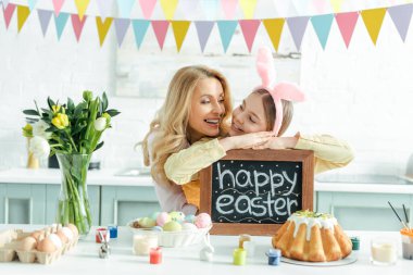 cheerful mother near daughter with bunny ears holding chalkboard with happy easter lettering 