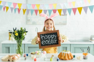 cheerful kid with bunny ears holding chalkboard with happy easter lettering 