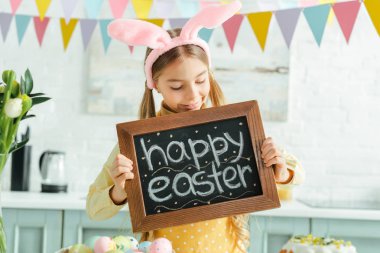 cheerful kid with bunny ears looking at chalkboard with happy easter lettering 