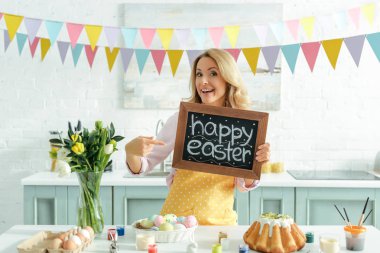 cheerful woman pointing with finger at chalkboard with happy easter lettering 