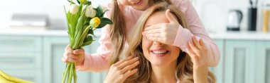 panoramic shot of kid covering eyes of happy mother while holding bouquet of tulips 