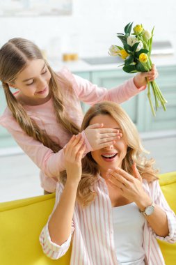 cute kid covering eyes of cheerful mother while holding bouquet of tulips 