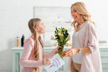 cheerful kid holding tulips and greeting card with happy mothers day lettering near mother 