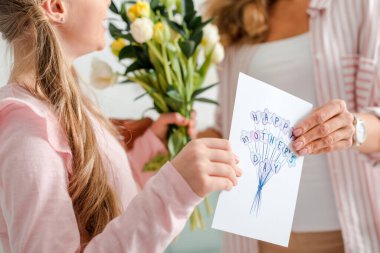 cropped view of happy kid holding tulips and greeting card with happy mothers day lettering near mother 