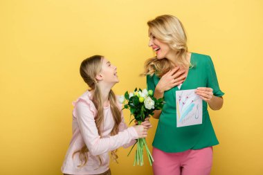 happy daughter giving tulips to mother holding greeting card with 8 march lettering isolated on yellow 