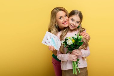 happy mother holding greeting card with 8 march lettering and hugging daughter with tulips isolated on yellow 