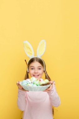 cheerful kid with bunny ears holding easter eggs in basket isolated on yellow 