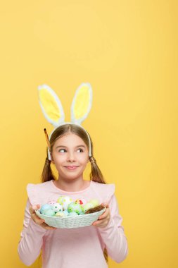 cheerful kid with bunny ears holding easter eggs in basket and looking away isolated on yellow 
