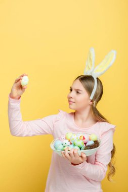 cute kid with bunny ears looking at easter egg isolated on yellow 