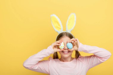 happy child with bunny ears covering eyes with easter eggs isolated on yellow 