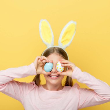 cheerful child with bunny ears covering eyes with easter eggs isolated on yellow 