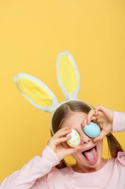 kid with bunny ears sticking out tongue and covering eyes with easter eggs isolated on yellow 