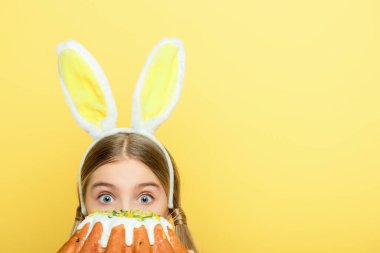 shocked kid with bunny ears covering face with easter cake isolated on yellow 