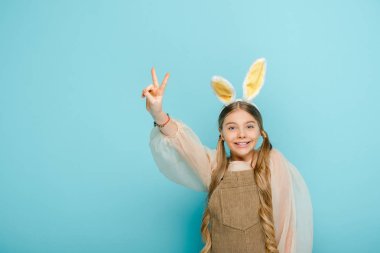 happy kid with bunny ears showing peace sign isolated on blue 