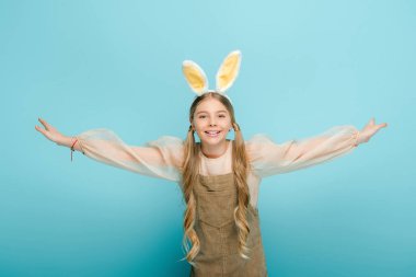 positive kid with bunny ears and outstretched hands isolated on blue 