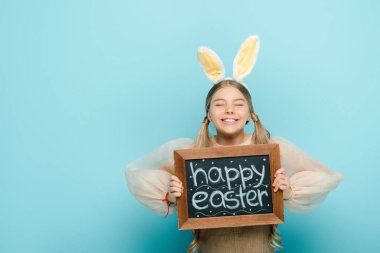 smiling kid with bunny ears holding chalkboard with happy easter lettering on blue 