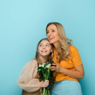happy mother and daughter smiling near tulips isolated on blue