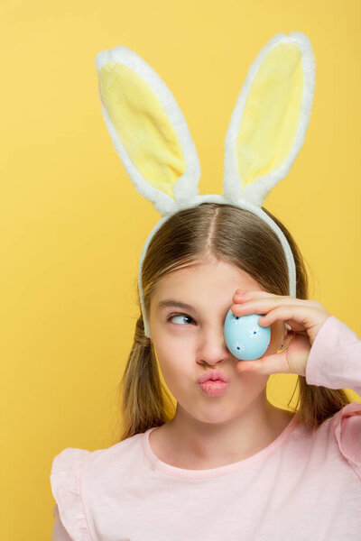 funny kid with bunny ears covering eye with dotted easter eggs isolated on yellow 