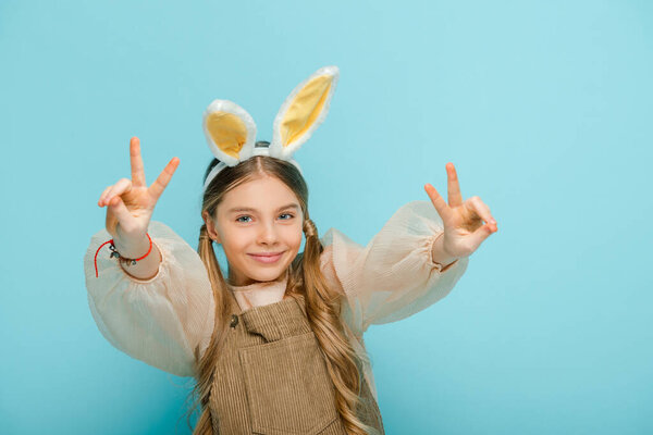 cheerful kid with bunny ears showing peace sign isolated on blue 