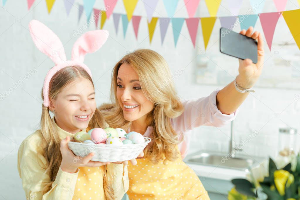 selective focus of happy mother and daughter in bunny ears taking selfie with painted easter eggs in basket 