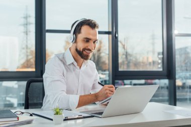positive translator working online with headphones and laptop in office clipart