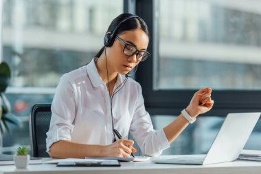 güzel Asyalı çevirmen ofiste kulaklık ve dizüstü bilgisayarla çevrimiçi çalışıyor