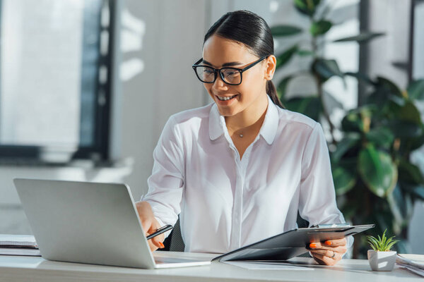 smiling asian translator in eyeglasses working online with laptop and documents