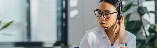 panoramic shot of asian translator in eyeglasses working online with headset and laptop in office