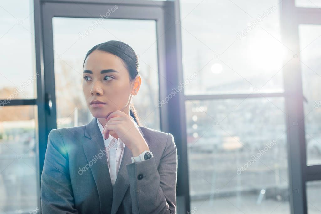 thoughtful asian businesswoman in grey suit standing in modern office