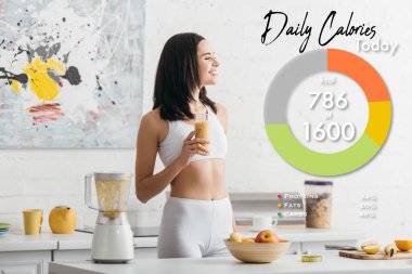Çekici fit sporcu kadın gülümsüyor ve elinde bir bardak smoothie ile mutfak masasının üzerinde günlük kalori resimleriyle ölçüm bandı tutuyor.