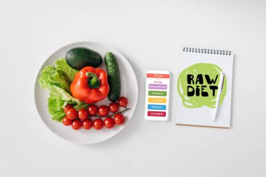 Top view of fresh vegetables and avocado on plate, smartphone and notebook on white background
