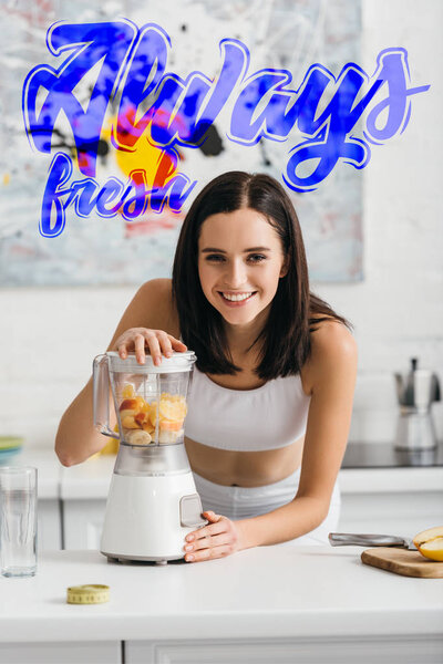 Selective focus of attractive sportswoman smiling at camera while preparing smoothie in blender near always fresh illustration