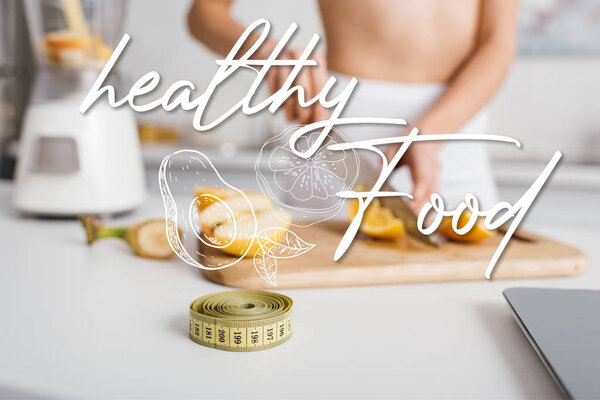 Селективный фокус измерения ленты и чешуи рядом с подходящей девушкой резки свежих фруктов для смузи на кухонном столе, иллюстрация здоровой пищи
