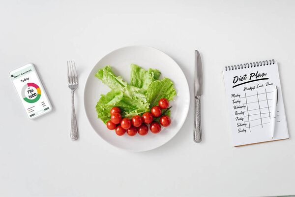 Top view of fresh vegetables on plate, smartphone with calorie counting app and notebook with diet plan on white background
