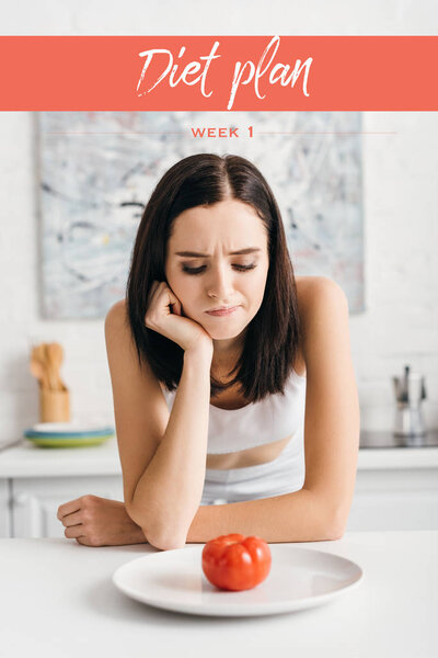 Pensive sportswoman looking at ripe tomato on plate on table near diet plan lettering