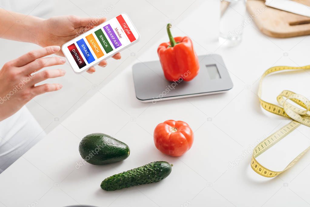 Cropped view of girl holding smartphone with daily diet plan app near vegetables, scales and measuring tape on kitchen table