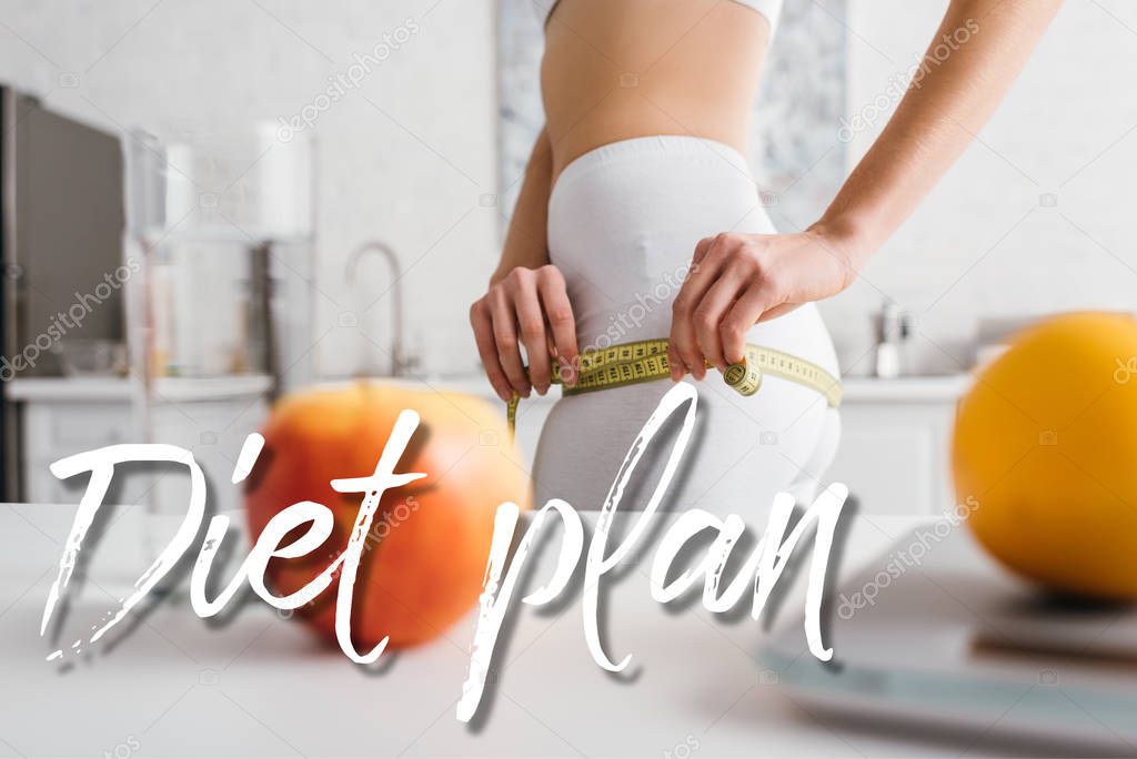 Selective focus of slim girl measuring hips near fruits, glass of water and scales on table in kitchen, diet plan illustration