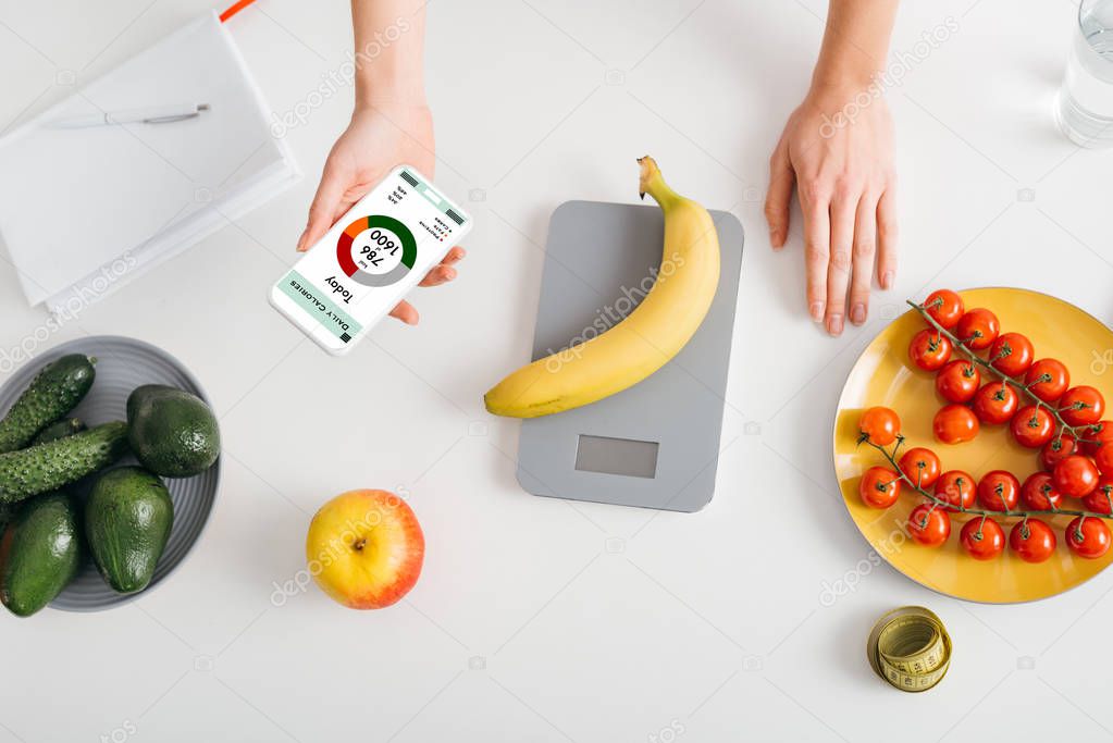 Top view of girl holding smartphone with calorie counting app while weighing banana on kitchen table near fresh vegetables