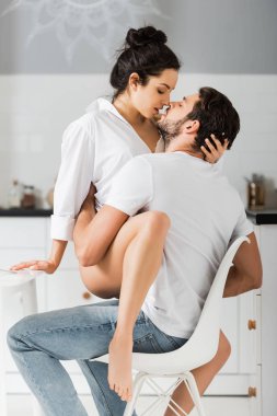 Sütyen ve gömlekli tutkulu bir kadın mutfakta sandalyede yakışıklı erkek arkadaşını öpüyor. 