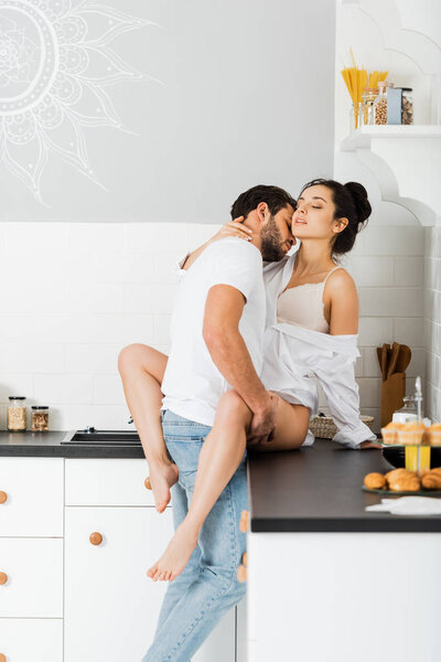 Handsome man kissing in neck and hugging seductive girlfriend in bra and shirt on kitchen worktop