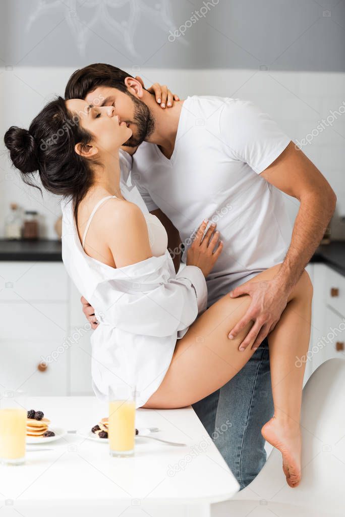 Side view of man kissing and touching by leg sensual woman in shirt and bra on table during breakfast in kitchen 