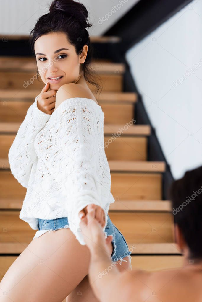 Selective focus of sensual woman holding hand of shirtless man on ladder at home
