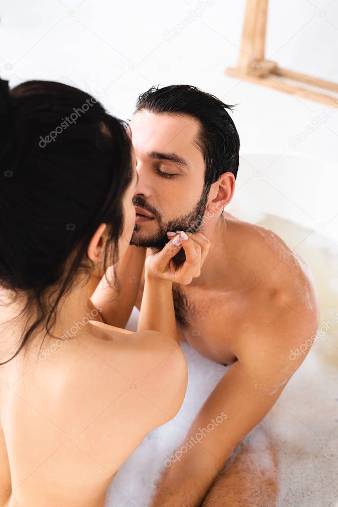 Selective focus of handsome man kissing girlfriend in bathtub with foam